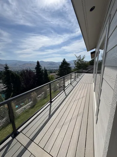 Long deck with pale boards and transparent glass railing overlooking trees and hills.