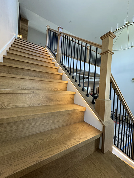 Wood staircase with black balusters, substantial newel posts and warm step lighting; floor protection remains in the adjacent room.