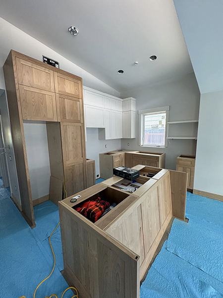 Natural wood kitchen cabinets and an island during installation over blue floor protection, view 3.