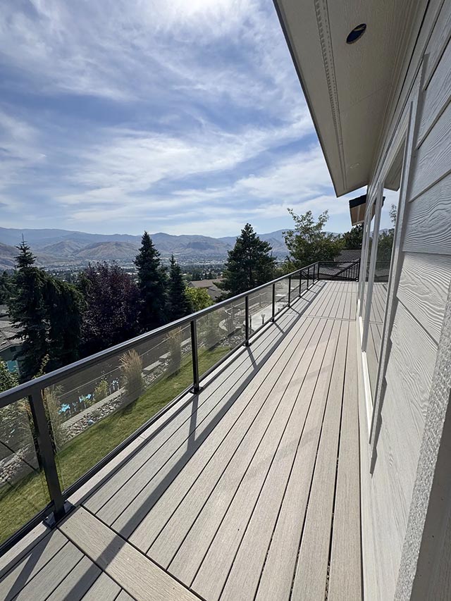 Long deck with pale boards and transparent glass railing overlooking trees and hills.